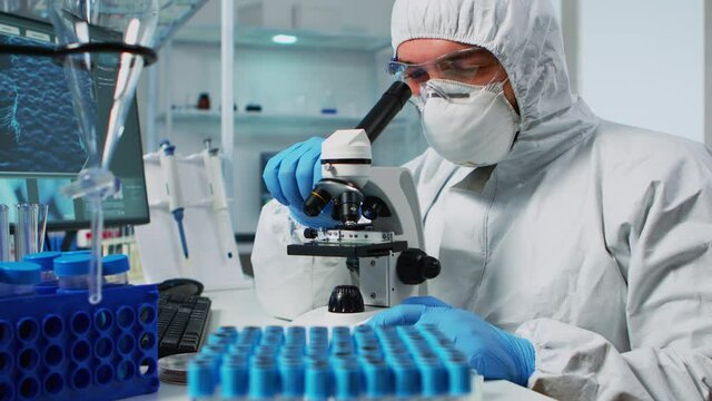 Biological scientist in coverall using microscope doing research for new vaccine in pharmaceutical labolatory. Coworkers examining virus evolution using high tech researching diagnosis against covid19