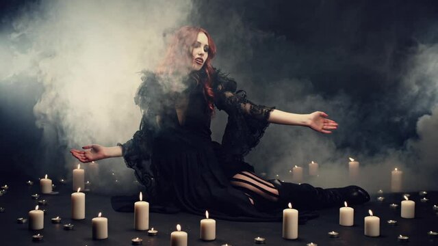 Red-haired witch sitting on floor in smoke, making energetic hand gestures to call demons. Magical ritual to invoke Satan. Cosplay, halloween costume. Concept of supernatural, evil, darkness, gothic.