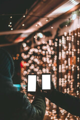 Two persons holding mobile phones in front of Christmas light bokeh. Blank screens for text or...