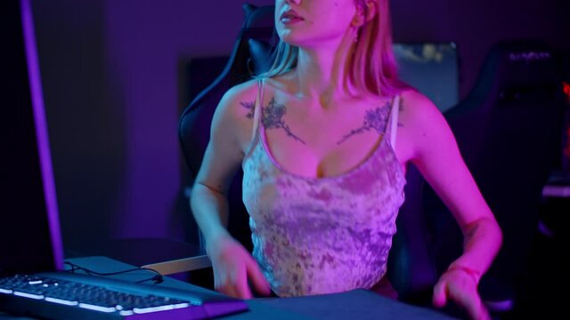 Tattooed gamer girl in sexy tank top sits by the PC - putting on her headphones and starts playing