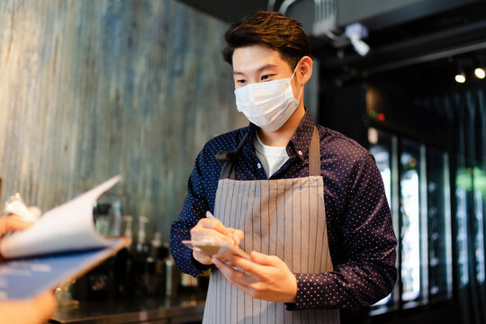 Asian Barista Man Wearing Mask Taking Order From Customer In Cafe.