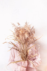 Pampas grass in woman's hands. Cortaderia selloana. Faceless woman in pink dress against abstract wall with champagne color dried flowers. 