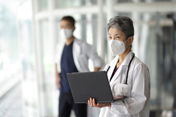 ノートパソコンを持つマスクをつけたシニア世代の日本人女性医師