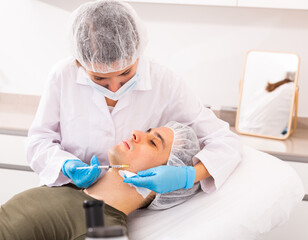 Fototapeta premium Experienced female beautician working with male client in clinic of aesthetic cosmetology, injecting facial filler into skin to correct face contour