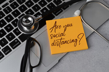 Selective focus of stethoscope, laptop, glasses and memo note written with question ARE YOU SOCIAL DISTANCING?. Health concept.