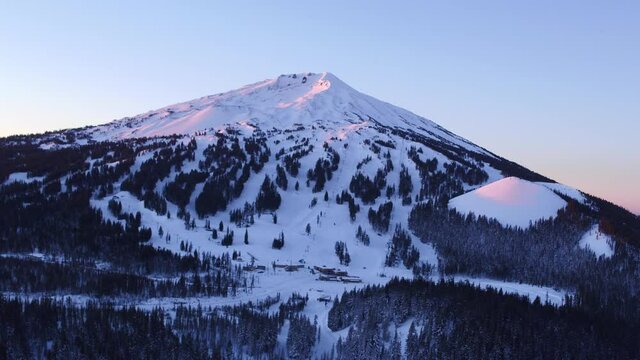 Aerial Horizontal drift (truck) across Mt Bachelor at Dawn/Sunrise on clear morning, soft blue & pink glow, trees covered with light snow, with an empty parking lot and no one skiing or snowboarding.
