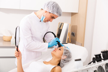 Young woman getting needle-free mesotherapy for face and looking in mirror during procedure