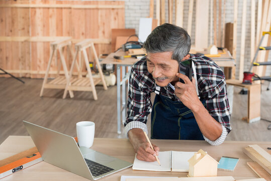 Senior asian elderly old man carpenter using mobile smartphone and laptop to contact customer for service in workshop