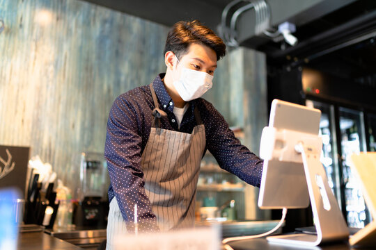 Asian barista man wearing mask working on digital menu in laptop.