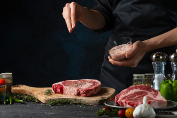 Macro shot of chef pours sea salt on raw steak on wooden chopped board. Backstage of preparing grilled pork meat at restaurant kitchen on dark blue background. Frozen motion. Cooking process.