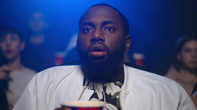 African man watching horror movie in cinema. Shocked afro guy breathing deep