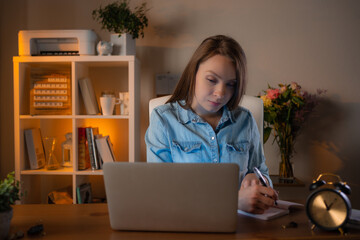 Young woman siting at her workplace in office, using laptop for searching information and makes notes in a notebook. Lovely job. Concept of working moments and office lifestyle.