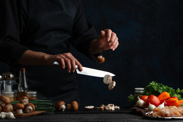 Professional chef in black uniform cuts with knife mushrooms on the chopped board. Concept of cooking process. Backstage of preparing meat filling for pie on dark blue background. Frozen motion.