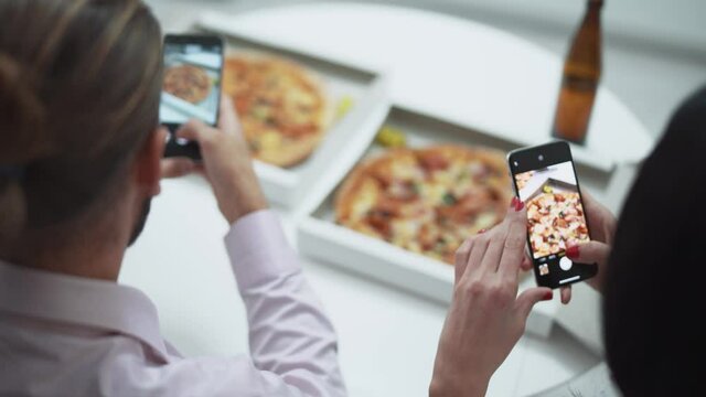 Home party, a young couple sitting at the table in the kitchen and taking pictures of pizza on smartphones.