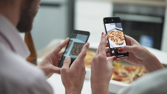 Home party, a young couple sitting at the table in the kitchen and taking pictures of pizza on smartphones.