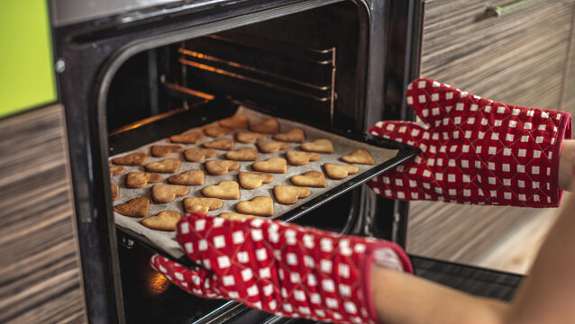 Woman is taking out a baking tray with delicious homemade heart-shaped cookies from the oven. Concept of fresh homemade cakes or a surprise for Valentine's Day - Powered by Adobe