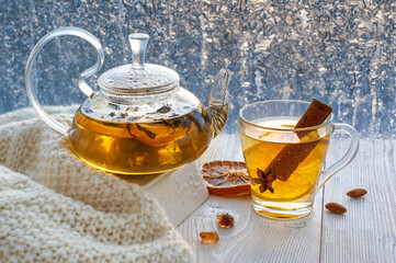 Winter warming tea with lemon and orange against the background of frosty patterns. Hot drink in a glass kettle and a cup on a wooden table in the background of the window.
