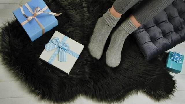 Female legs in warm socks rest comfortably on a gray fur skin next to blue and white gift boxes with bows. Expectation of the winter holidays Christmas and New Year. Close up. Slow motion.