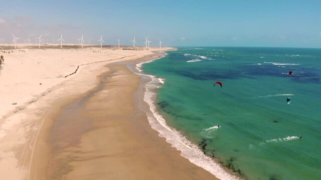 Drone view of kiteboarding, white sandy beach, turquoise ocean water, rustic fisherman boats and windmills in the background. Kitesurfing paradise near Taiba in Brazil.
