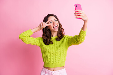 Photo of funky lady hold cellphone shoot selfie show v-sign near eye wear green sweater isolated pink color background