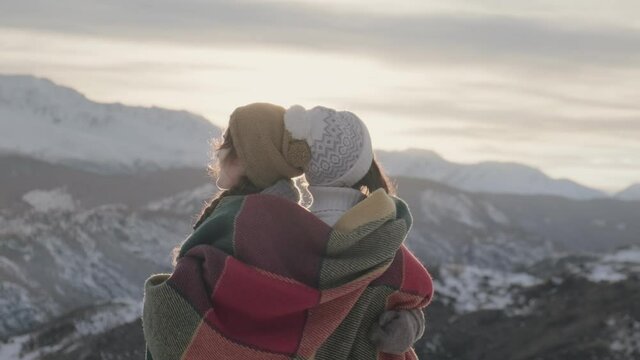 Two women wrap themselves in a woolen blanket to keep warm and enjoy the view of the setting sun over the mountains on a winter day