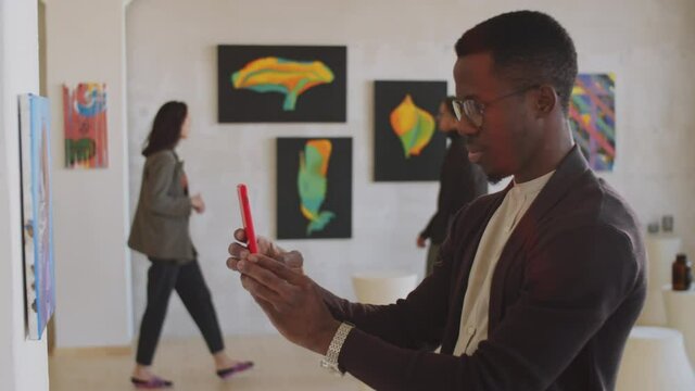 Side view of young Afro-American man taking picture of artwork with smartphone and smiling while visiting exhibition in gallery