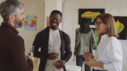 PAN shot of young multiethnic male and female guests holding champagne glasses, smiling and chatting at exhibition opening party in art gallery