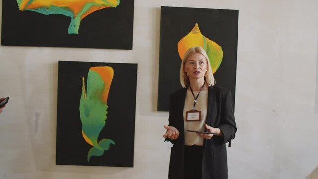 PAN shot of professional female guide holding digital tablet and speaking about paintings on the wall with visitors while working at modern art exhibition in gallery
