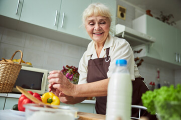 Cheerful old woman in apron cooking dinner at home