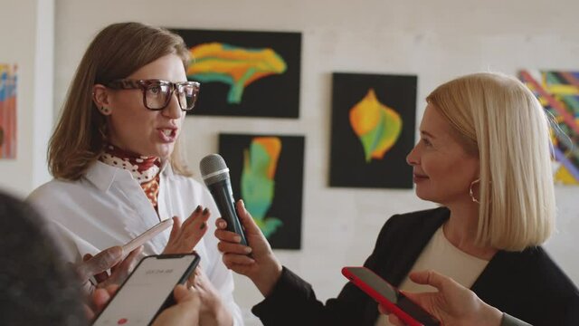 Young female artist smiling and talking to journalists while giving interview at art exhibition opening in gallery