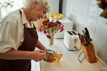 Cheerful old woman pouring herbal tea into cup in kitchen