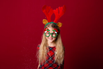 cute little girl in Christmas glasses blows snow from palms in the studio on a red background. Christmas concept, text space