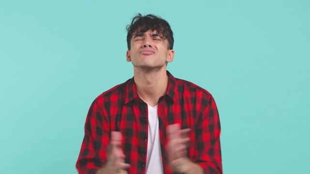 Dissatisfied crying young man 20s in basic red checkered shirt posing isolated on blue turquoise color background studio. People lifestyle concept. Spreading arms hold hands folded in prayer ask why