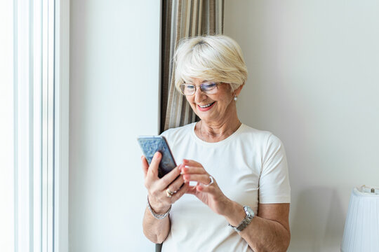 Portrait Of Cheerful Senior Woman Using Smartphone Woman Relaxing At Home Reading Her Text Messages On Her Mobile Phone With A Quiet Smile. Senior Female Texting Or Playing An Online Game