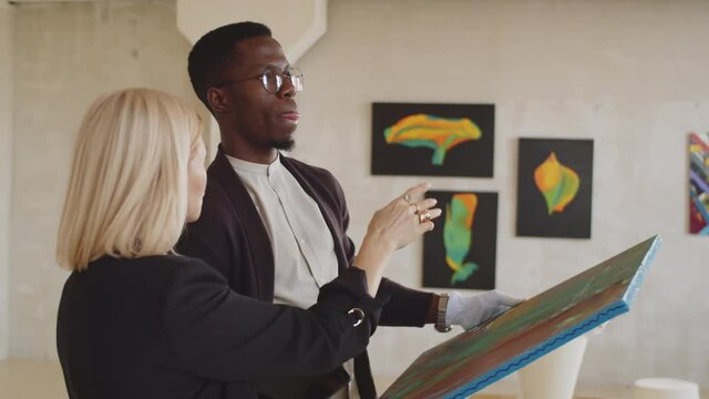 Middle aged Caucasian female manager telling where to hang painting to young black male worker in white gloves while preparing exhibition in art gallery