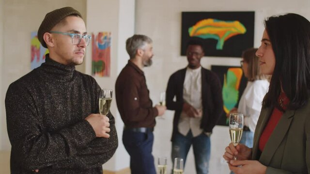 PAN waist up shot of young man and woman holding champagne glasses and chatting at exhibition opening party in art gallery