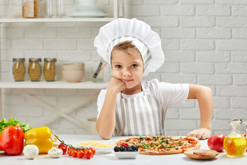 cute little boy in chef hat and an apron cooking pizza in the kitchen. child looking at the camera