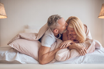 Grey-haired man and a blonde woman lying on the bed and looking romantic