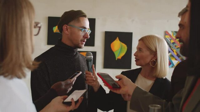 Young male artist giving interview to group of multiethnic journalists at art exhibition opening in gallery
