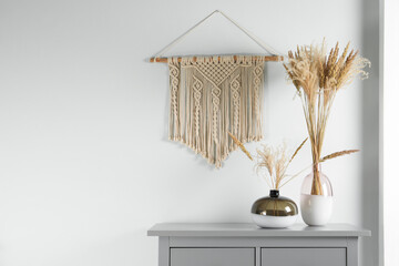 Reed's blossom in glass vases on grey cabinet near white wall with decorative element. Space for...
