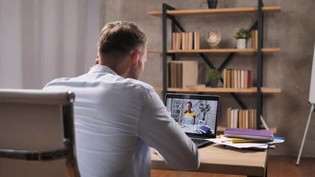 Rear view of young man meeting remotely with mixed race female interior designer during discussion of apartment renovation. Customer of redevelpment project conversating on laptop with woman designer