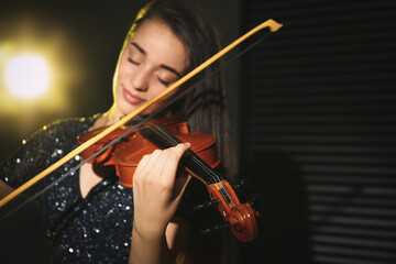 Fototapeta premium Beautiful young woman playing violin in dark room, focus on hand
