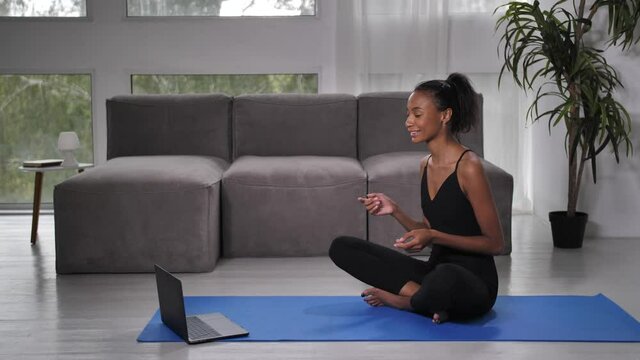 Positive fit female instructor conversating with client using video chat on laptop while sitting in yoga lotus pose. Joyful mixed race online coach teaching yoga practices and meditation at distance