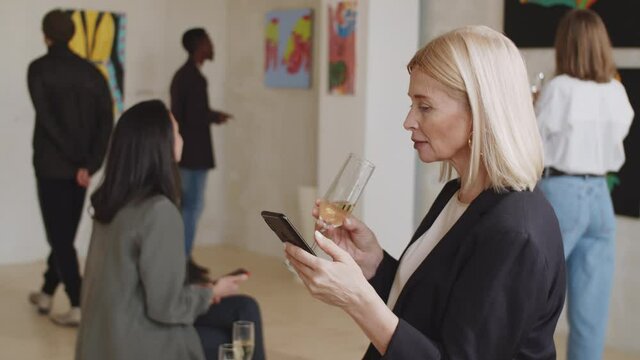 Beautiful mid-aged lady drinking champagne and typing on smartphone in art gallery while visitors watching exhibition