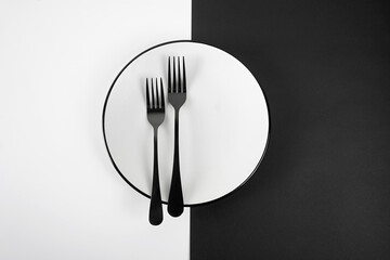 Two black fork on the white plate. Two parts background: black and white. Minimalist. Meniu concept. Flat lay.