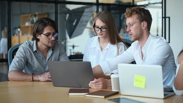 Modern young business people in glasses sitting by the table and working and using laptops together.