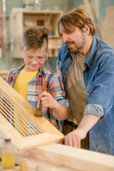 Fototapeta na wymiar Father teaches his young son how to paint a wooden board with a paintbrush in a carpentry workshop