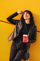 Young hipster girl in a black jacket with a red paper cup of coffee on a yellow background with a...