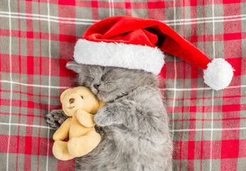 Obraz na płótnie Canvas Cute kitten wearing red santa's hat sleeps on checkered or tartan blanket blanket and hugs favorite toy bear