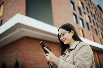 Beautiful young woman with eyeglasses standing on the street near brick building and is using smartphone. 
